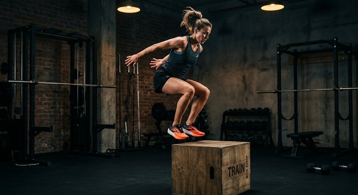 An athlete wearing Brooks Hyperion 3 trainers performing a dynamic movement in a moody, professional gymnasium setting.