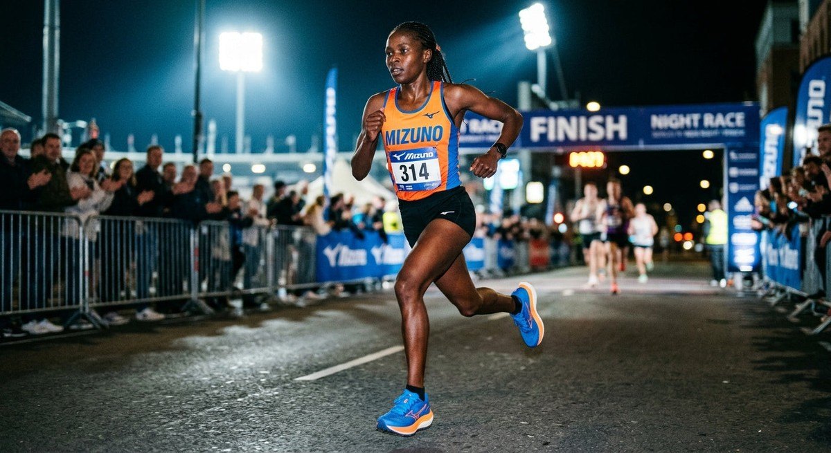 An elite athlete wearing the Mizuno Hyperwarp mid-stride during a night race, capturing the sharp details of the monofilament mesh upper and stadium atmosphere.