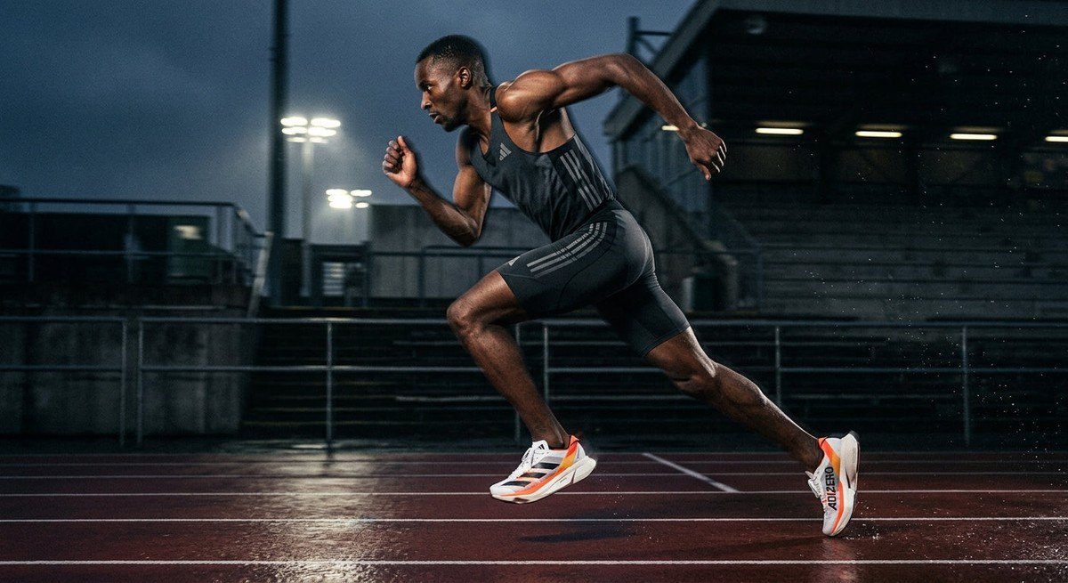 Athletic side profile of a runner in motion blur, showcasing the aggressive geometric rocker profile of the new Adidas racing shoe in a moody stadium environment.