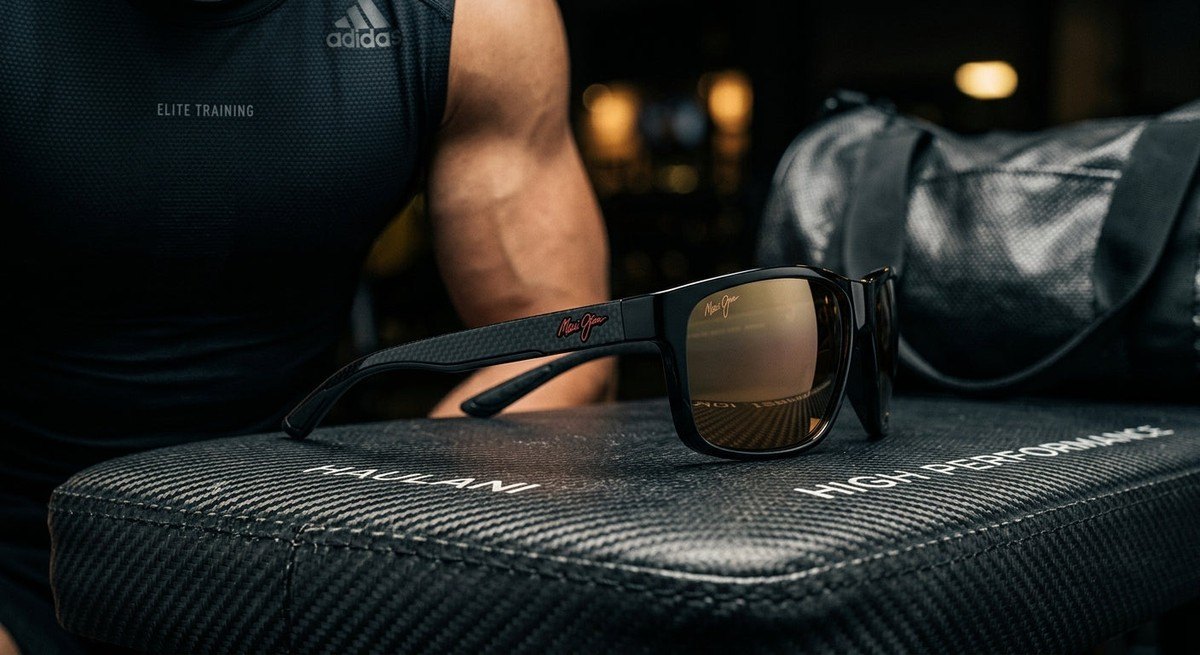 Cinematic side profile of the Maui Jim Haulani sunglasses resting on a carbon fibre surface with dramatic studio lighting.