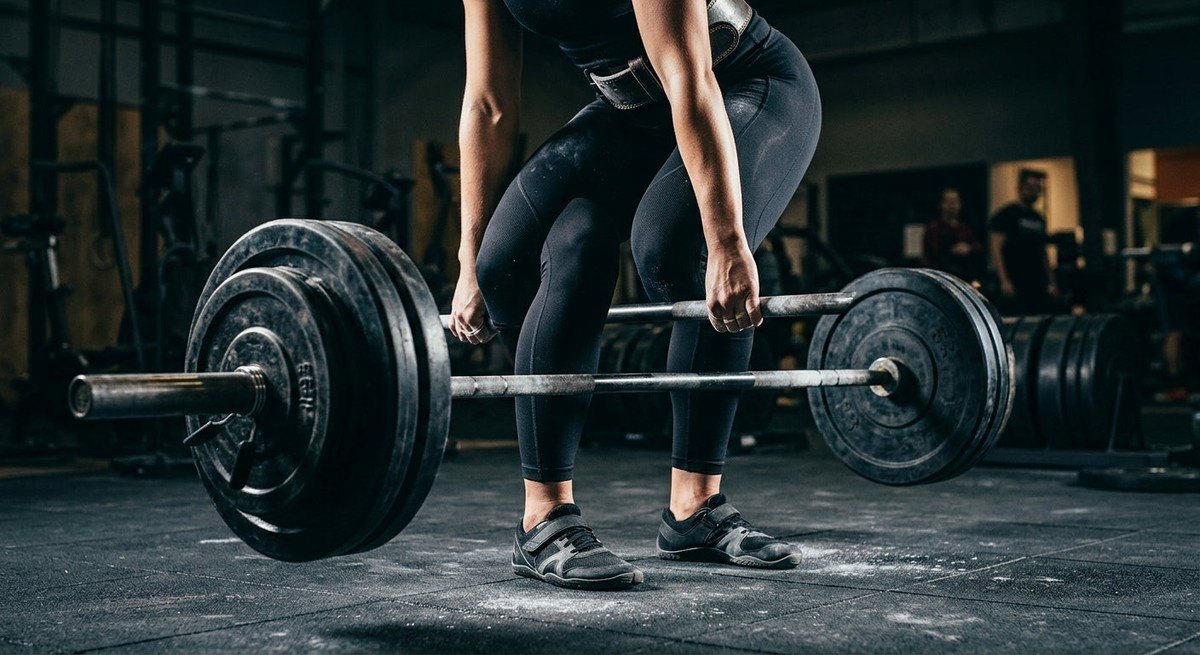 Close-up action shot of a runner's legs during a heavy deadlift, capturing the intensity of motion and the fine textures of gym equipment with a dark, moody background.