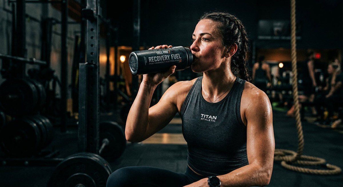Close-up of a high-performance athlete drinking a recovery supplement bottle in a moody, high-contrast gym environment.