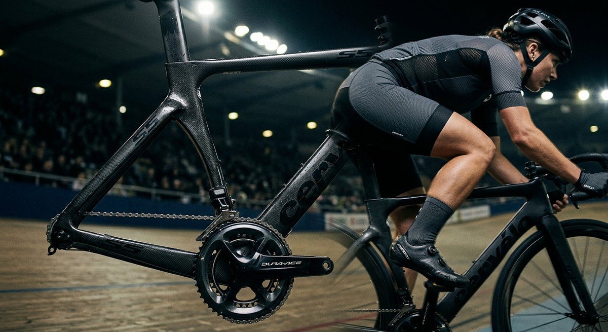 Close-up of a high-performance carbon fibre road bike frame under moody stadium lights, emphasising texture and sharp mechanical details.