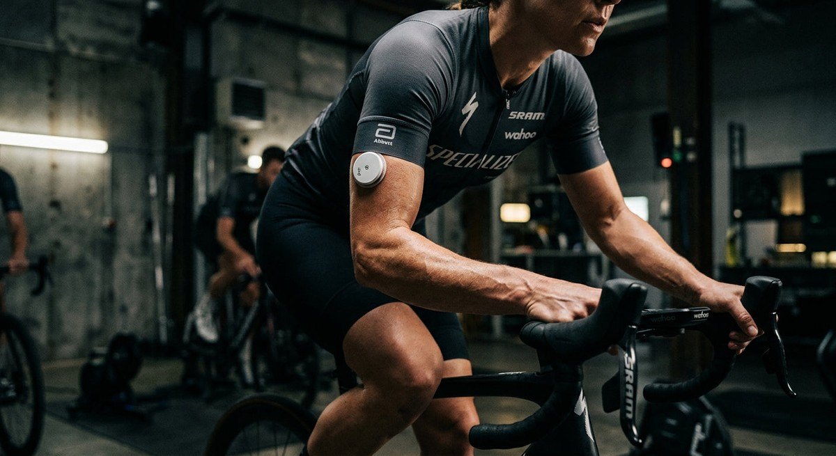 Close-up of a high-tech glucose sensor on a cyclist's arm, with the background showing a high-contrast, moody training environment.