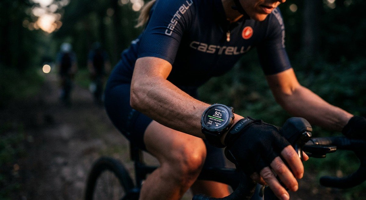 Close-up of a high-tech wearable fitness tracker on a cyclist's wrist, displaying recovery data in a moody, low-light environment.