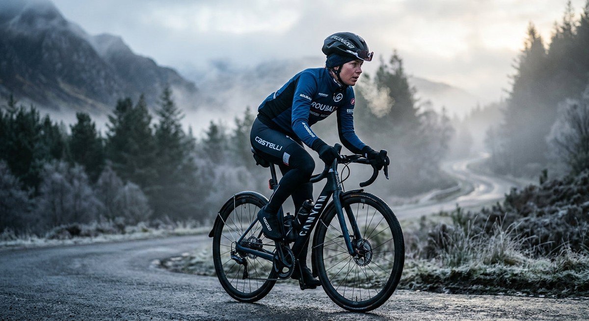 Close-up of a road cyclist wearing professional winter gear riding through a misty, atmospheric landscape during a winter morning.