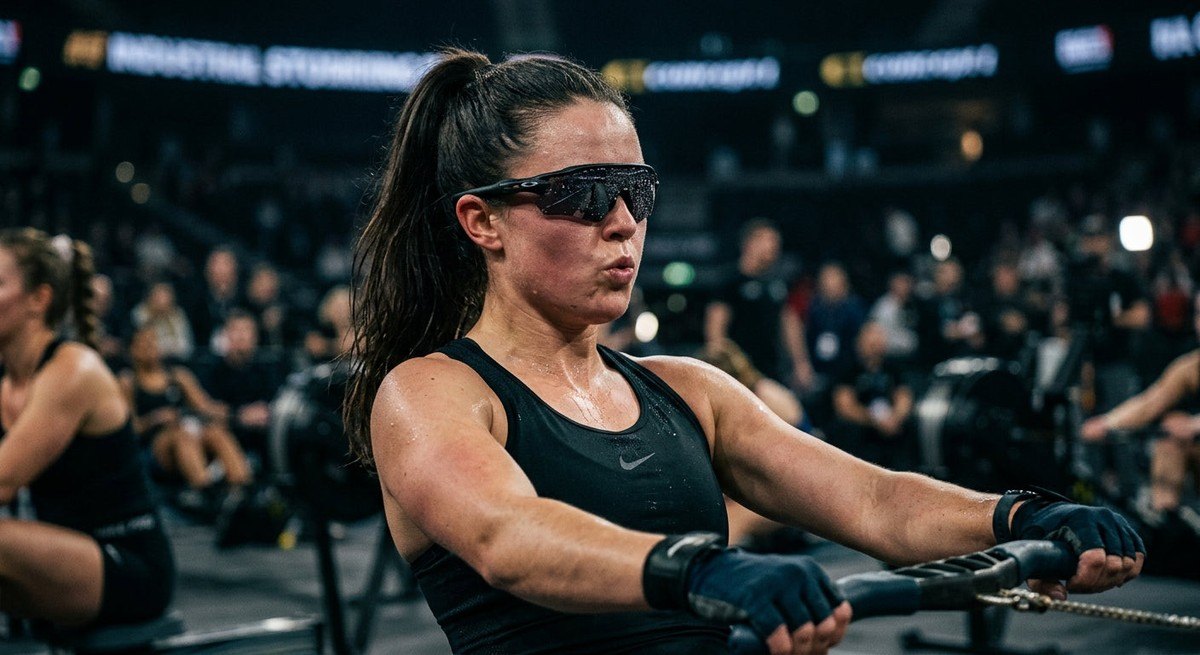 Close-up of a rower's face wearing sleek wrap-around sports sunglasses, water droplets on the lens, moody stadium lighting background.