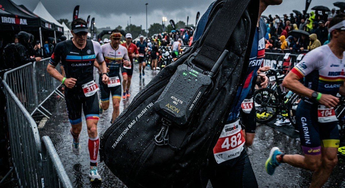 Close-up of a sleek, ruggedised UWB tracking device mounted securely to a transition bag, with motion-blurred athletes rushing through a rainy, high-density triathlon race environment.