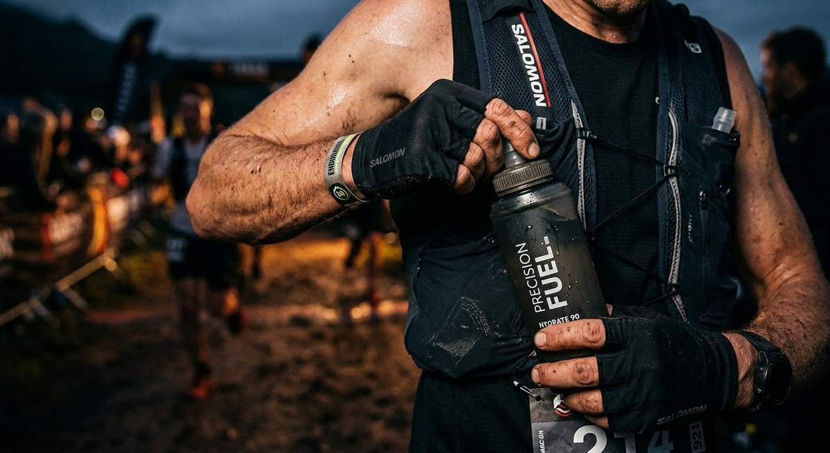 Close-up of an athlete's hands adjusting a precision fuel flask during a race, sharp textures, moody lighting, and professional athletic gear in focus.