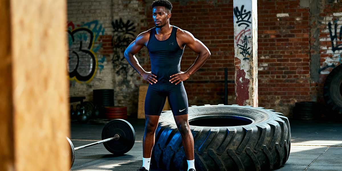 A full length shot of a Black male athlete standing next to a gym tyre in a modern training facility.