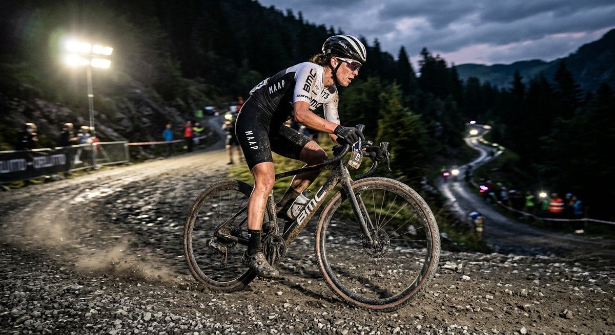 Professional cyclist in high-speed action, navigating a technical, winding gravel ascent with dramatic stadium-style lighting illuminating the grit.