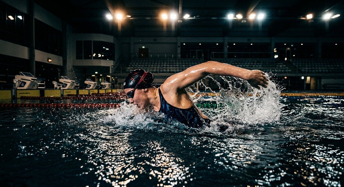 Professional swimmer captured in a dramatic, high-contrast action shot during an intense training session with realistic water textures and stadium lights